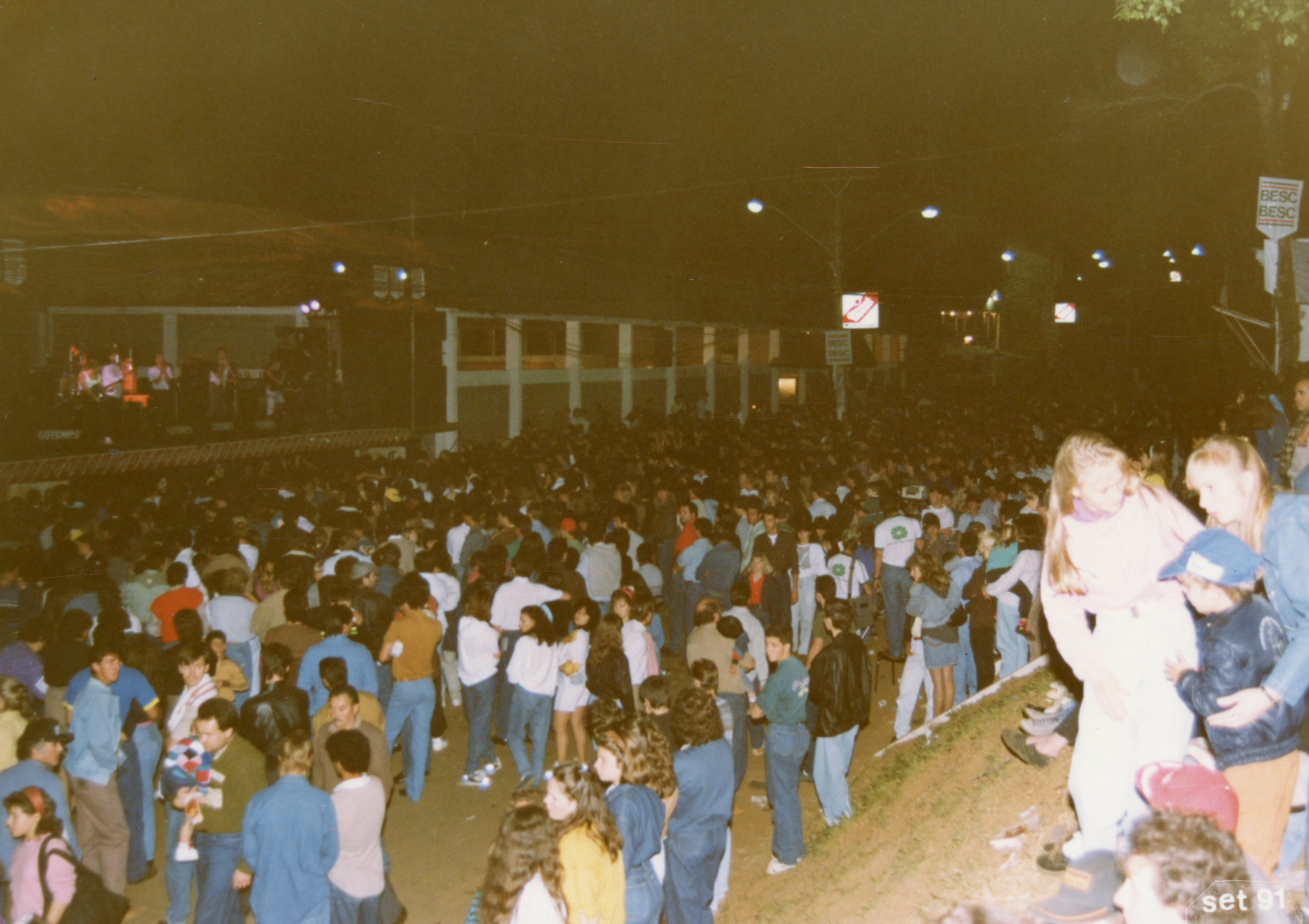 Uma fotografia noturna de um evento público ao ar livre, possivelmente um show ou festival, com uma grande multidão reunida em frente a um palco iluminado. A imagem apresenta vestimentas típicas do início dos anos 90 e a presença de sinalização do banco BESC, sugerindo um contexto de evento comunitário em uma cidade brasileira.