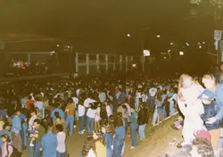 Uma fotografia noturna de um evento público ao ar livre, possivelmente um show ou festival, com uma grande multidão reunida em frente a um palco iluminado. A imagem apresenta vestimentas típicas do início dos anos 90 e a presença de sinalização do banco BESC, sugerindo um contexto de evento comunitário em uma cidade brasileira.