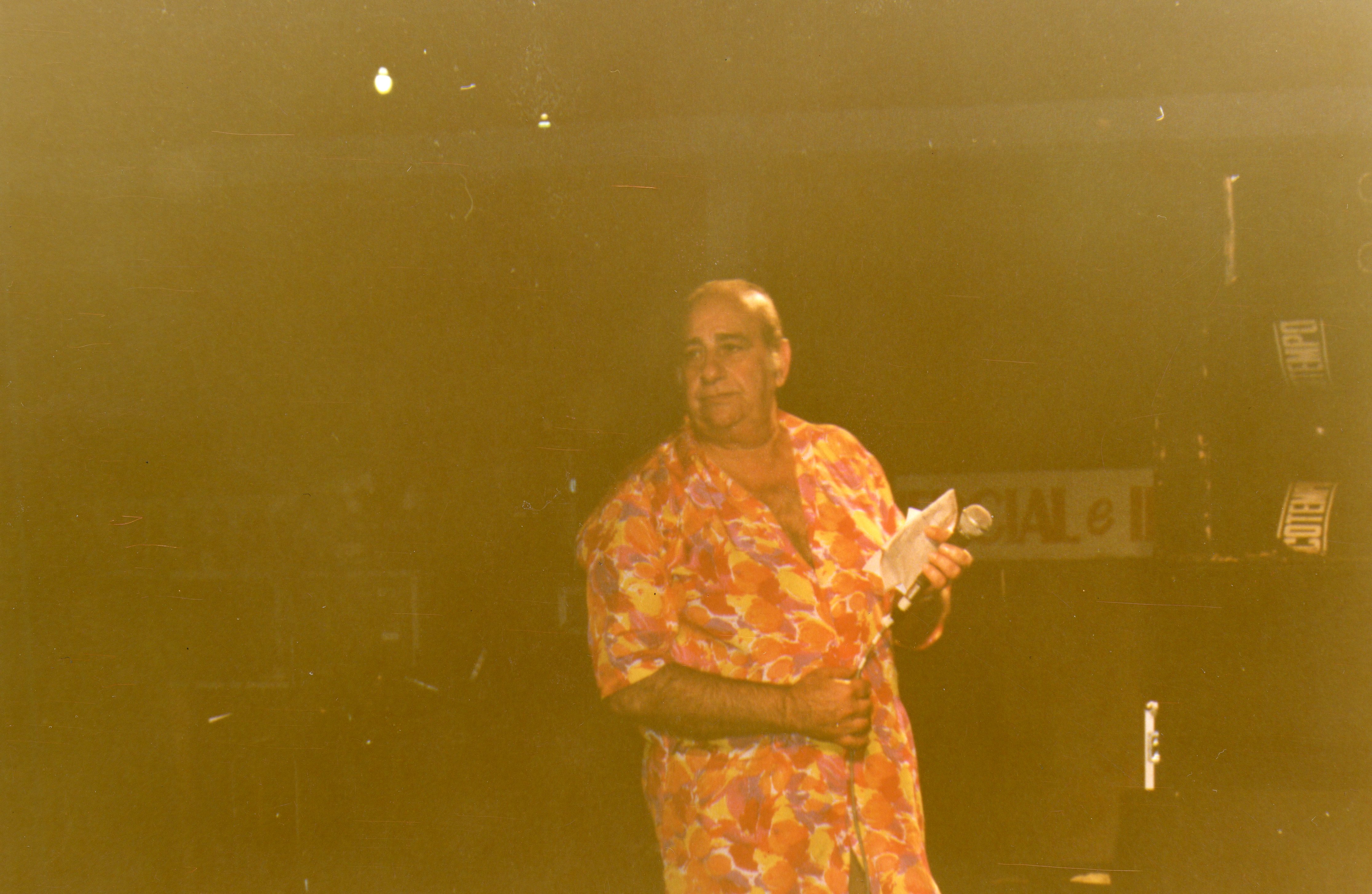 Uma fotografia de um homem de meia-idade vestindo uma camisa estampada colorida, segurando um microfone e um papel, possivelmente em um evento ou apresentação pública. A imagem possui um tom sépia característico de fotografias amadoras das décadas de 1970 ou 1980, sugerindo um ambiente de entretenimento ou celebração local.