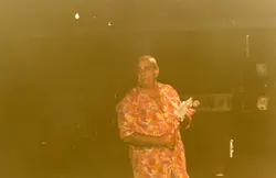 Uma fotografia de um homem de meia-idade vestindo uma camisa estampada colorida, segurando um microfone e um papel, possivelmente em um evento ou apresentação pública. A imagem possui um tom sépia característico de fotografias amadoras das décadas de 1970 ou 1980, sugerindo um ambiente de entretenimento ou celebração local.