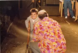 Uma fotografia que captura um momento de interação em um palco, possivelmente durante um evento de entretenimento ou show. Um homem de baixa estatura, vestindo um terno claro e usando uma muleta, sorri para um homem de costas que veste uma camisa estampada colorida, com uma plateia visível ao fundo em um ambiente de baixa luminosidade.