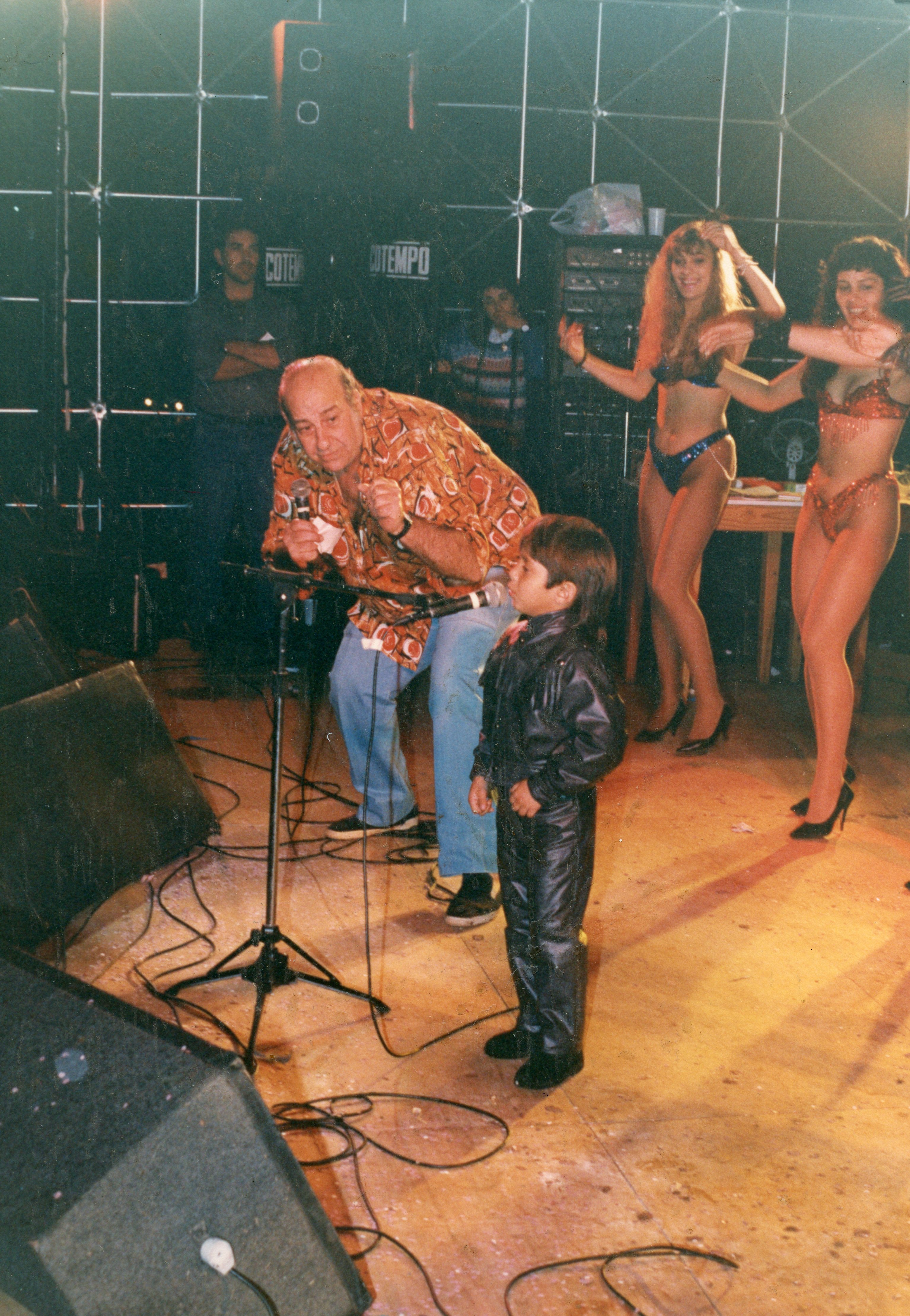 Uma fotografia de um evento de entretenimento, possivelmente um show ou programa de auditório, apresentando um homem adulto com microfone e uma criança vestida de couro ao lado. Ao fundo, duas mulheres com trajes de palco dançam, sugerindo uma atmosfera de espetáculo popular típica das décadas de 1980 ou 1990.