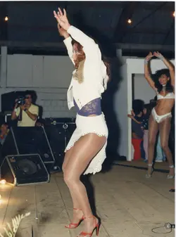 Uma fotografia que captura uma apresentação de dança em um ambiente interno, possivelmente um clube ou salão de eventos. As dançarinas vestem trajes típicos de espetáculos de palco dos anos 80 ou 90, enquanto um fotógrafo registra o momento ao fundo.