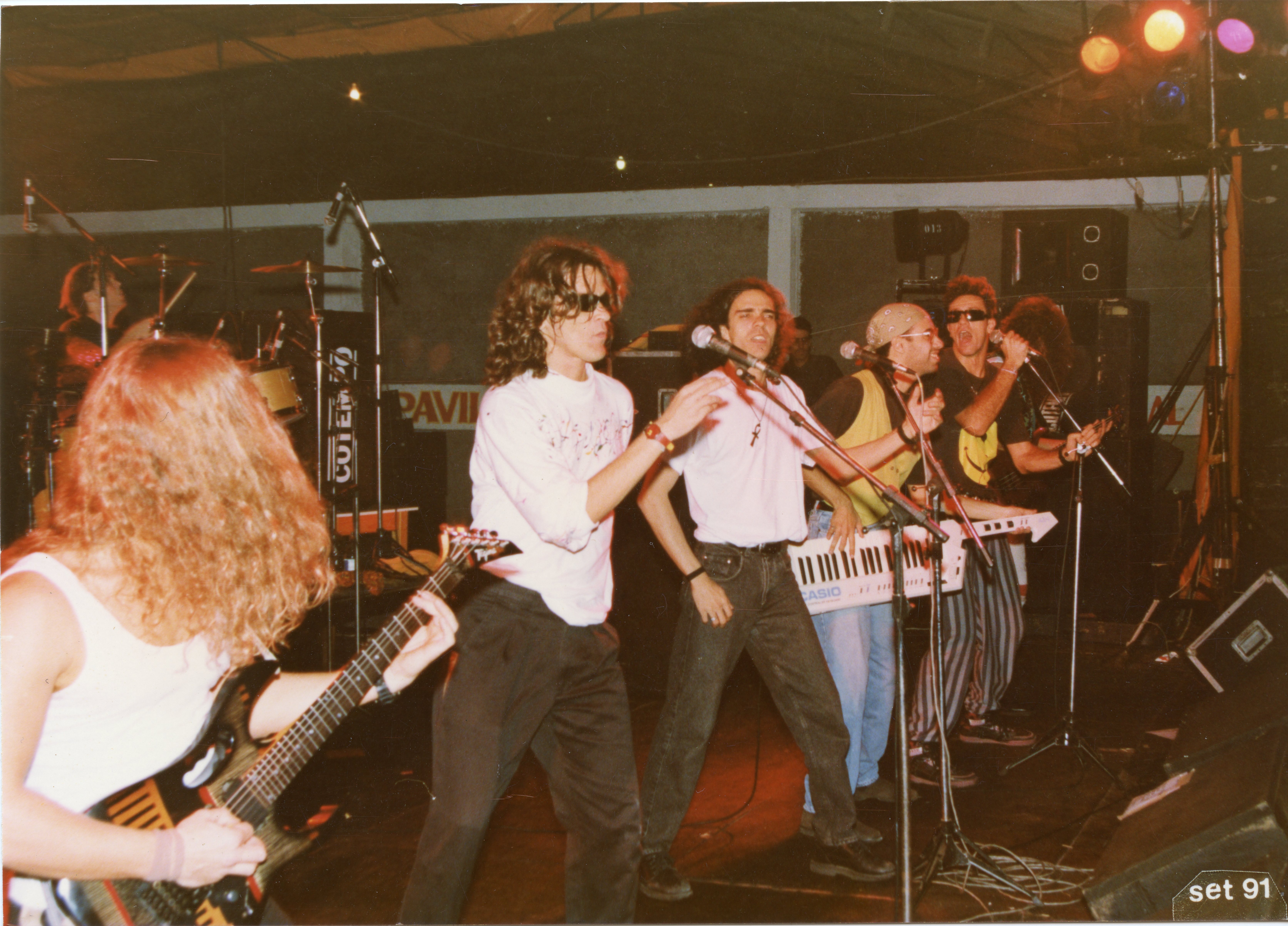 Uma banda de rock brasileira se apresentando ao vivo em um palco coberto, com integrantes utilizando instrumentos típicos da época, como um teclado keytar. A estética e o vestuário, incluindo óculos escuros e camisetas, remetem ao cenário musical do início da década de 1990.