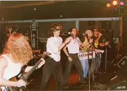 Uma banda de rock brasileira se apresentando ao vivo em um palco coberto, com integrantes utilizando instrumentos típicos da época, como um teclado keytar. A estética e o vestuário, incluindo óculos escuros e camisetas, remetem ao cenário musical do início da década de 1990.