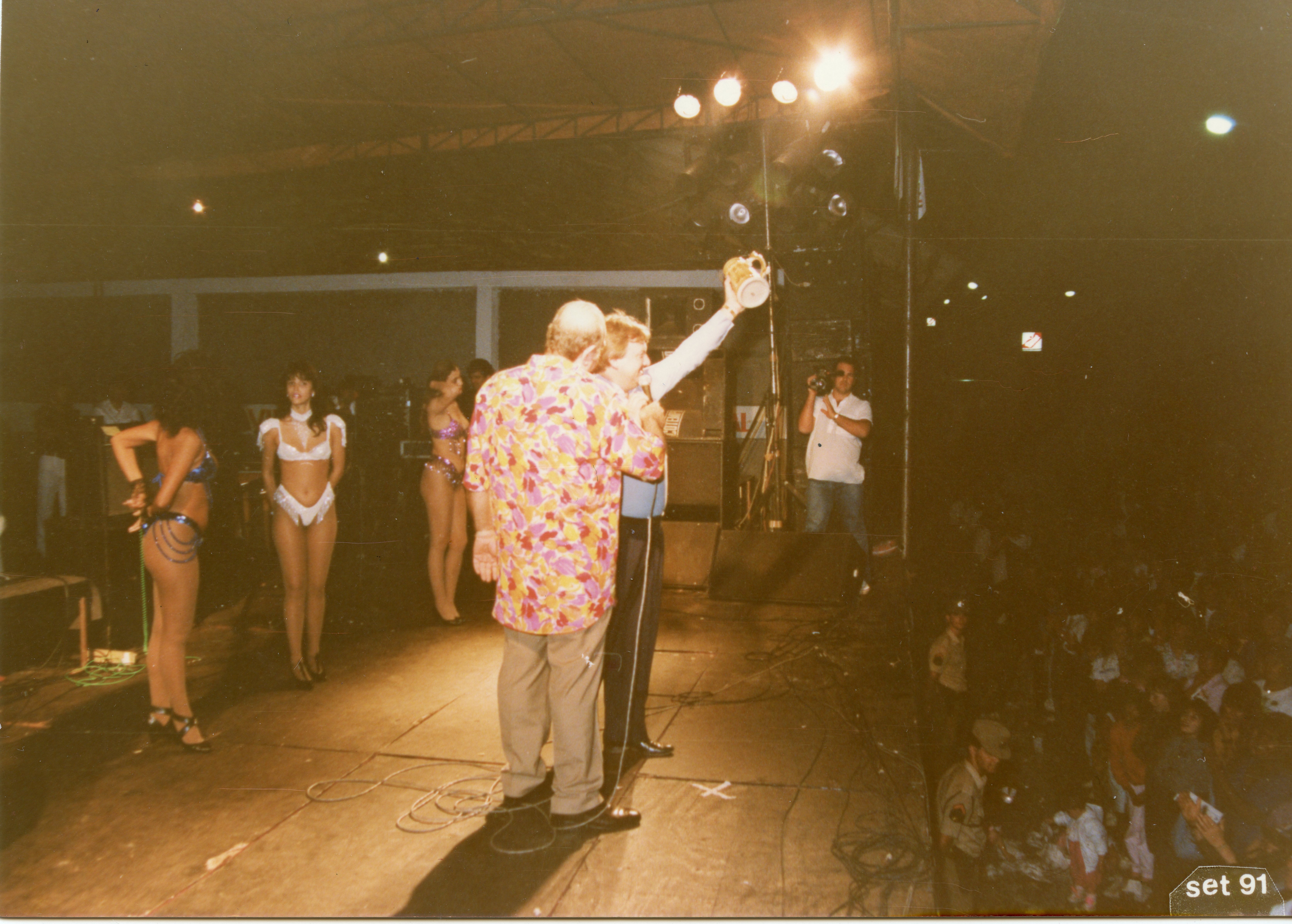 Uma fotografia noturna capturando um evento festivo em um palco, possivelmente uma celebração cultural ou festival de chopp. Vê-se um apresentador segurando uma caneca de chopp, acompanhado por mulheres vestidas com trajes de passista ou fantasia, sob iluminação de palco em um ambiente que sugere uma estrutura temporária.