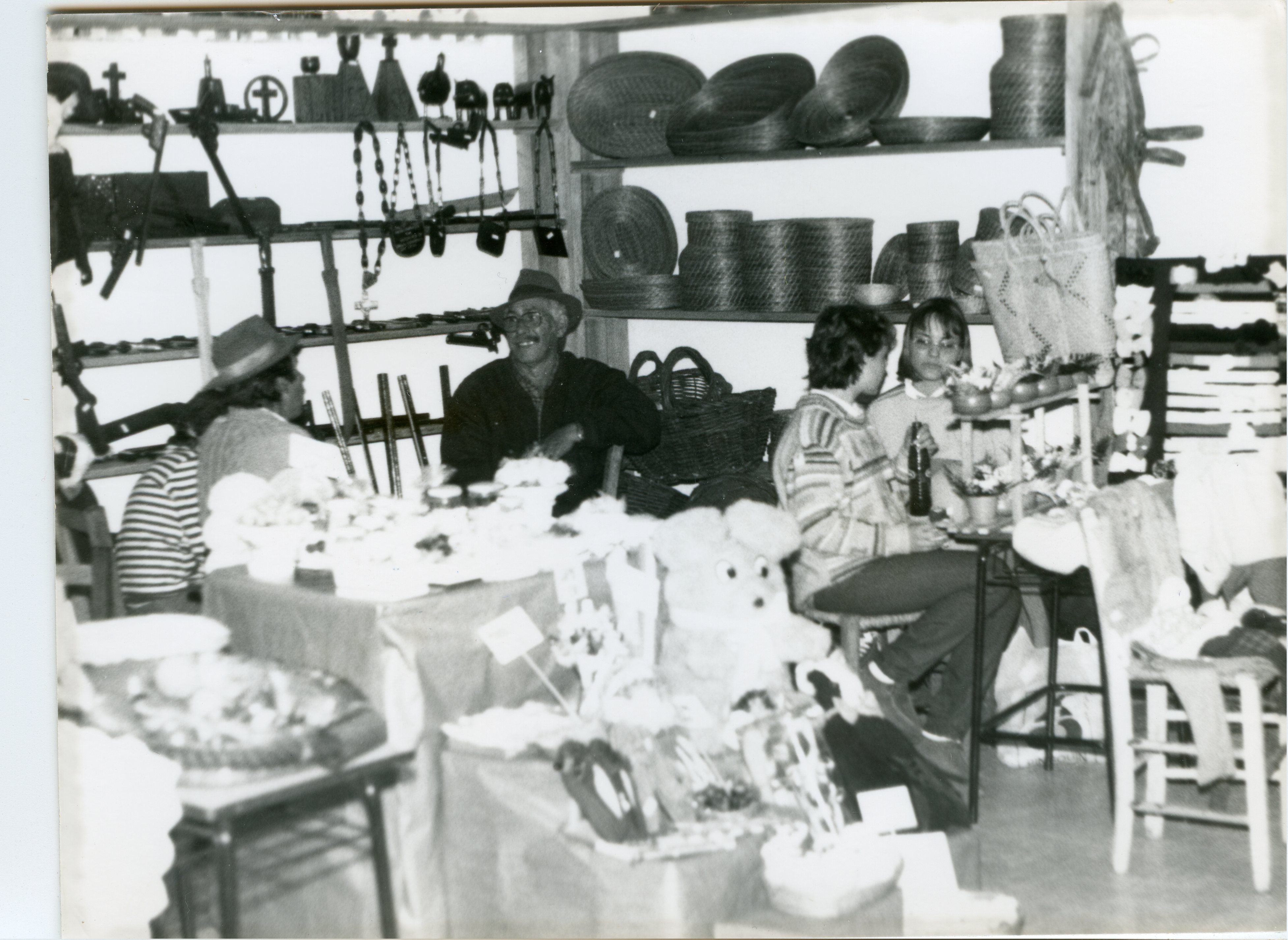 Uma fotografia em preto e branco que retrata uma feira de artesanato ou exposição cultural, possivelmente em um contexto regional brasileiro. Vê-se pessoas sentadas atrás de mesas com produtos artesanais, como cestarias e objetos de madeira, sugerindo um evento comunitário ou de valorização da cultura local.