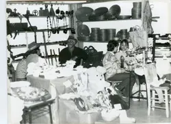 Uma fotografia em preto e branco que retrata uma feira de artesanato ou exposição cultural, possivelmente em um contexto regional brasileiro. Vê-se pessoas sentadas atrás de mesas com produtos artesanais, como cestarias e objetos de madeira, sugerindo um evento comunitário ou de valorização da cultura local.