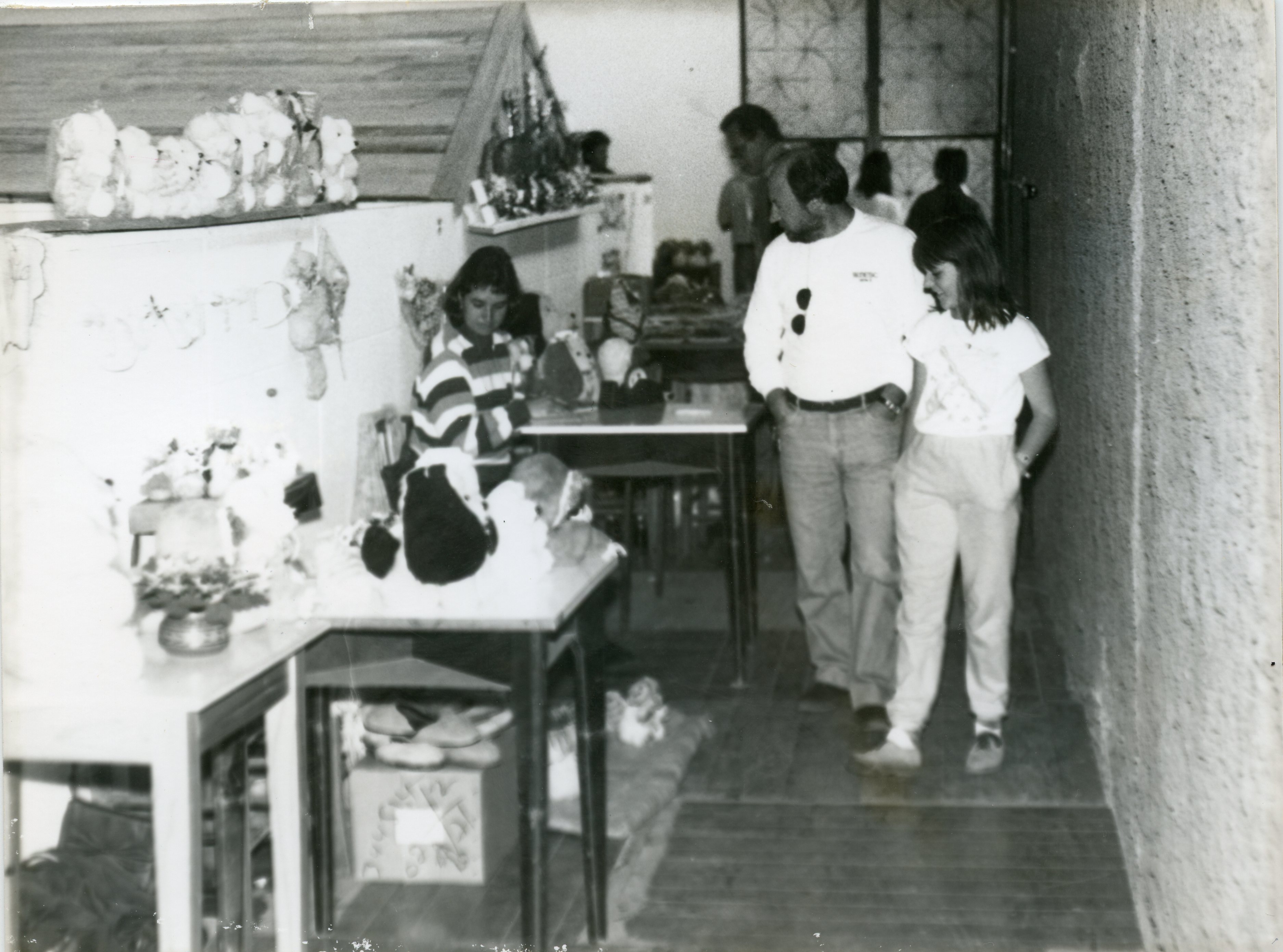 Fotografia em preto e branco registrando um ambiente interno, possivelmente uma feira de artesanato ou exposição escolar, com mesas contendo objetos decorativos e bonecos. Um homem e uma jovem caminham pelo corredor lateral, enquanto outra pessoa está sentada ao fundo, sugerindo um evento comunitário típico das décadas de 1980 ou 1990.