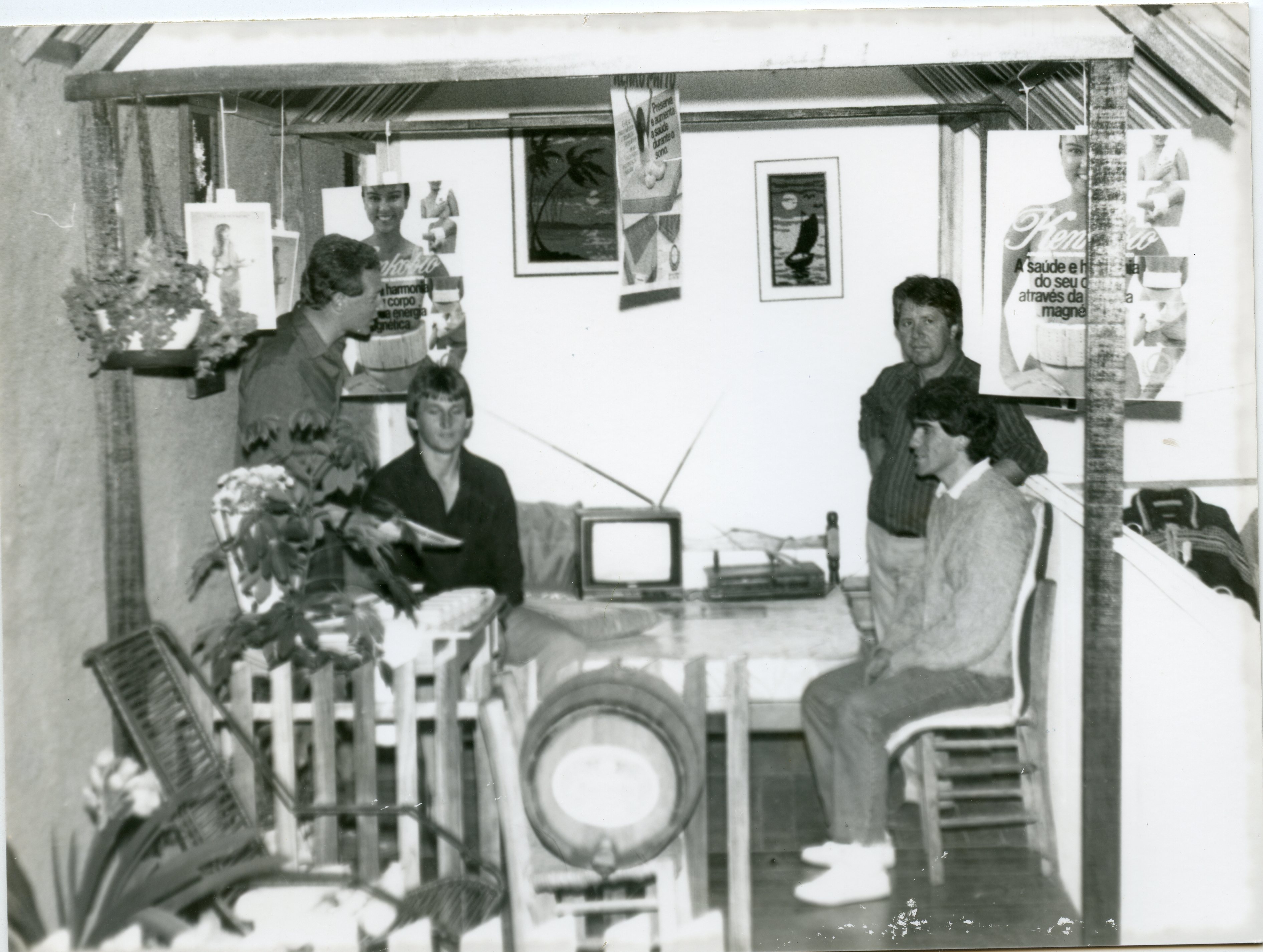 Uma fotografia em preto e branco que mostra quatro homens reunidos em um ambiente que parece ser um estande de exposição ou uma pequena estrutura comercial improvisada. O local é decorado com cartazes publicitários de produtos de saúde e bem-estar, sugerindo uma feira ou evento promocional, possivelmente ocorrido entre as décadas de 1980 e 1990.