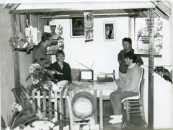 Uma fotografia em preto e branco que mostra quatro homens reunidos em um ambiente que parece ser um estande de exposição ou uma pequena estrutura comercial improvisada. O local é decorado com cartazes publicitários de produtos de saúde e bem-estar, sugerindo uma feira ou evento promocional, possivelmente ocorrido entre as décadas de 1980 e 1990.
