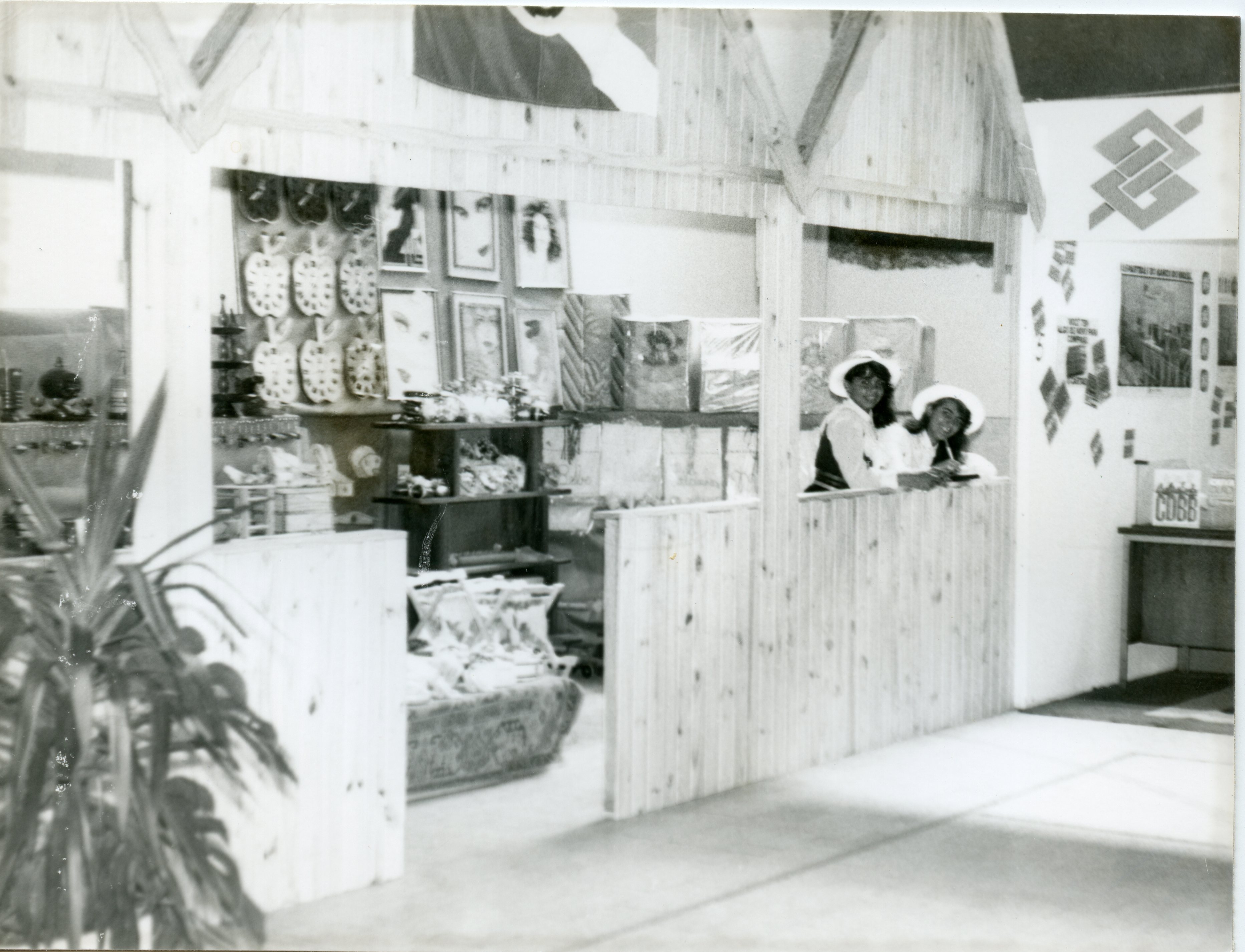 Fotografia em preto e branco mostrando um estande de exposição, possivelmente em uma feira comercial ou agrícola, com duas mulheres usando chapéus atrás de um balcão de madeira. O ambiente apresenta elementos decorativos como quadros e objetos em formato de maçã, além do logotipo do Banco do Brasil na parede, sugerindo um contexto de promoção institucional ou regional.