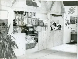 Fotografia em preto e branco mostrando um estande de exposição, possivelmente em uma feira comercial ou agrícola, com duas mulheres usando chapéus atrás de um balcão de madeira. O ambiente apresenta elementos decorativos como quadros e objetos em formato de maçã, além do logotipo do Banco do Brasil na parede, sugerindo um contexto de promoção institucional ou regional.