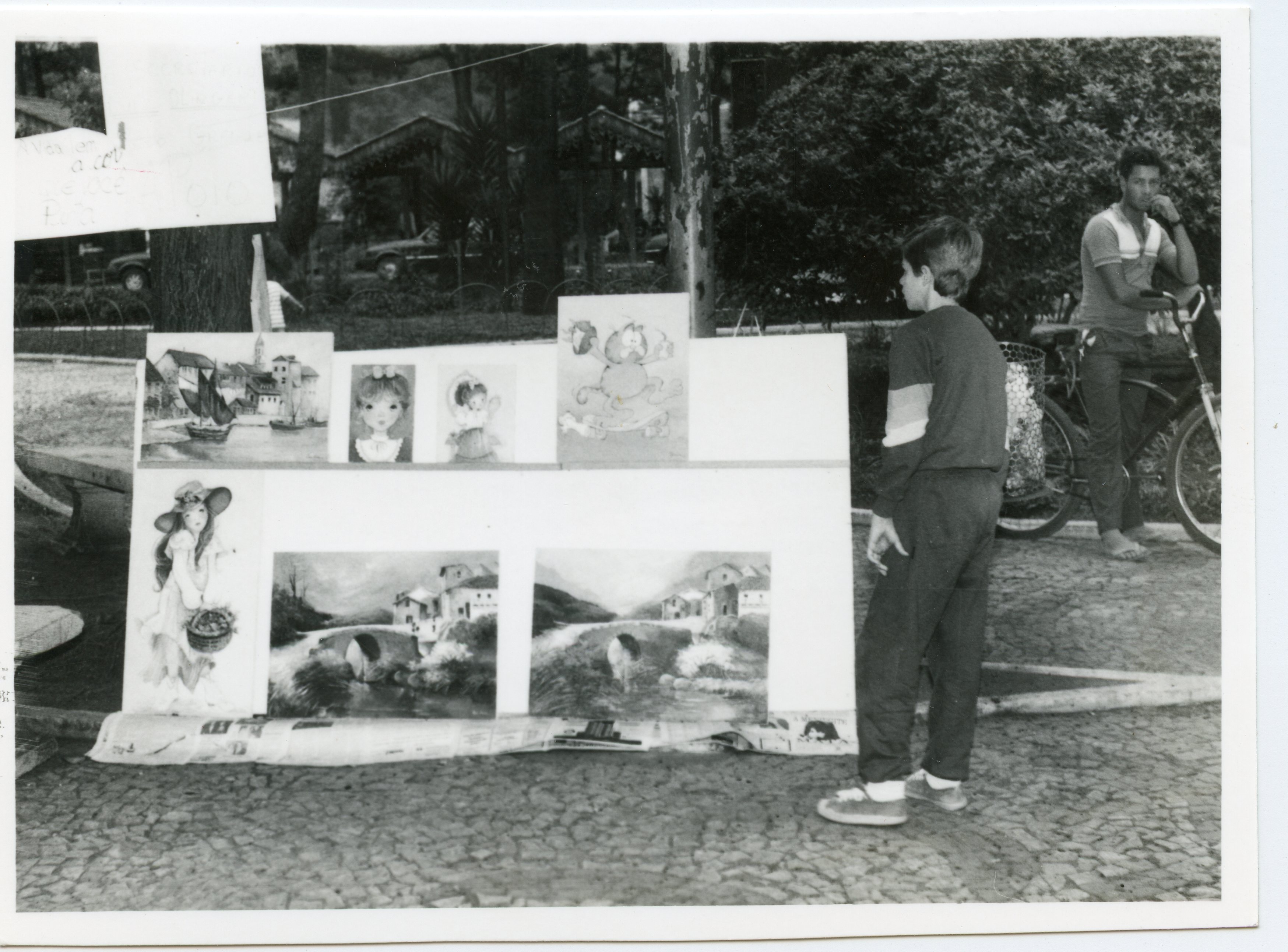 Uma fotografia em preto e branco que retrata uma exposição de quadros ao ar livre em uma praça, possivelmente em uma cidade brasileira. Um jovem observa as pinturas enquanto um homem adulto posa ao lado de uma bicicleta, sugerindo uma cena cotidiana de lazer ou comércio informal típica das décadas de 1980 ou 1990.