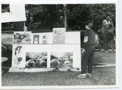 Uma fotografia em preto e branco que retrata uma exposição de quadros ao ar livre em uma praça, possivelmente em uma cidade brasileira. Um jovem observa as pinturas enquanto um homem adulto posa ao lado de uma bicicleta, sugerindo uma cena cotidiana de lazer ou comércio informal típica das décadas de 1980 ou 1990.