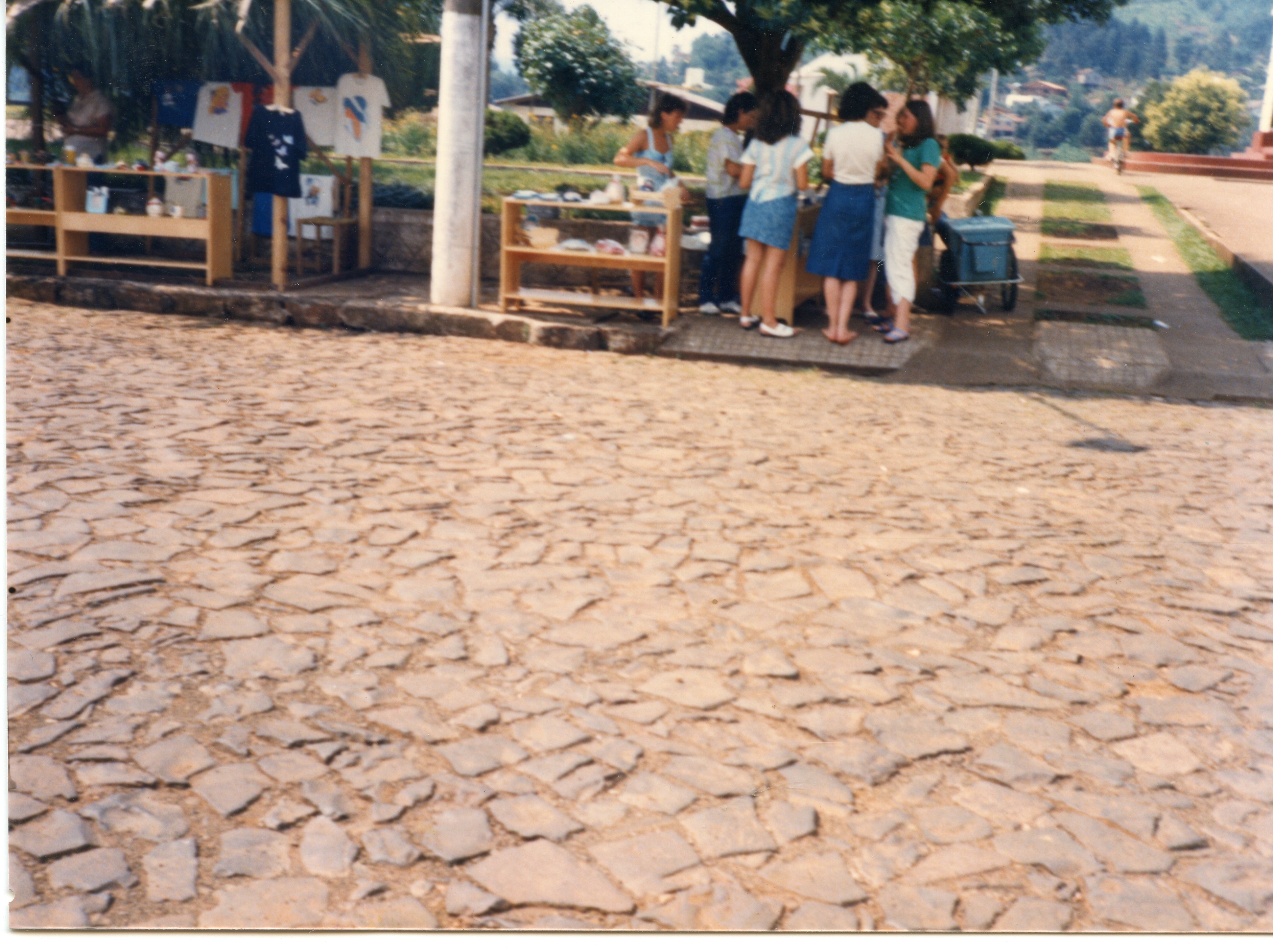 Uma cena ao ar livre mostrando um pequeno grupo de pessoas reunidas em torno de bancas de artesanato ou venda de roupas em uma praça pavimentada com pedras irregulares. O estilo das vestimentas, como saias e blusas, sugere uma atmosfera típica de uma cidade do interior brasileiro, possivelmente durante a década de 1980 ou início de 1990.