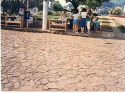 Uma cena ao ar livre mostrando um pequeno grupo de pessoas reunidas em torno de bancas de artesanato ou venda de roupas em uma praça pavimentada com pedras irregulares. O estilo das vestimentas, como saias e blusas, sugere uma atmosfera típica de uma cidade do interior brasileiro, possivelmente durante a década de 1980 ou início de 1990.