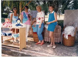 Uma fotografia colorida que mostra quatro mulheres em uma feira de artesanato ao ar livre, possivelmente em uma praça ou parque. Elas estão exibindo bonecas de pano e peças de crochê, com vestimentas típicas da década de 1990, sugerindo um evento comunitário ou cultural em uma cidade brasileira.