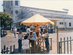 Uma cena ao ar livre em uma praça, possivelmente em frente a uma igreja, com pessoas interagindo em uma barraca de venda de brinquedos de pelúcia sob um guarda-sol amarelo. A vestimenta e a qualidade da fotografia sugerem um cenário brasileiro típico das décadas de 1980 ou 1990, com arquitetura religiosa ao fundo.