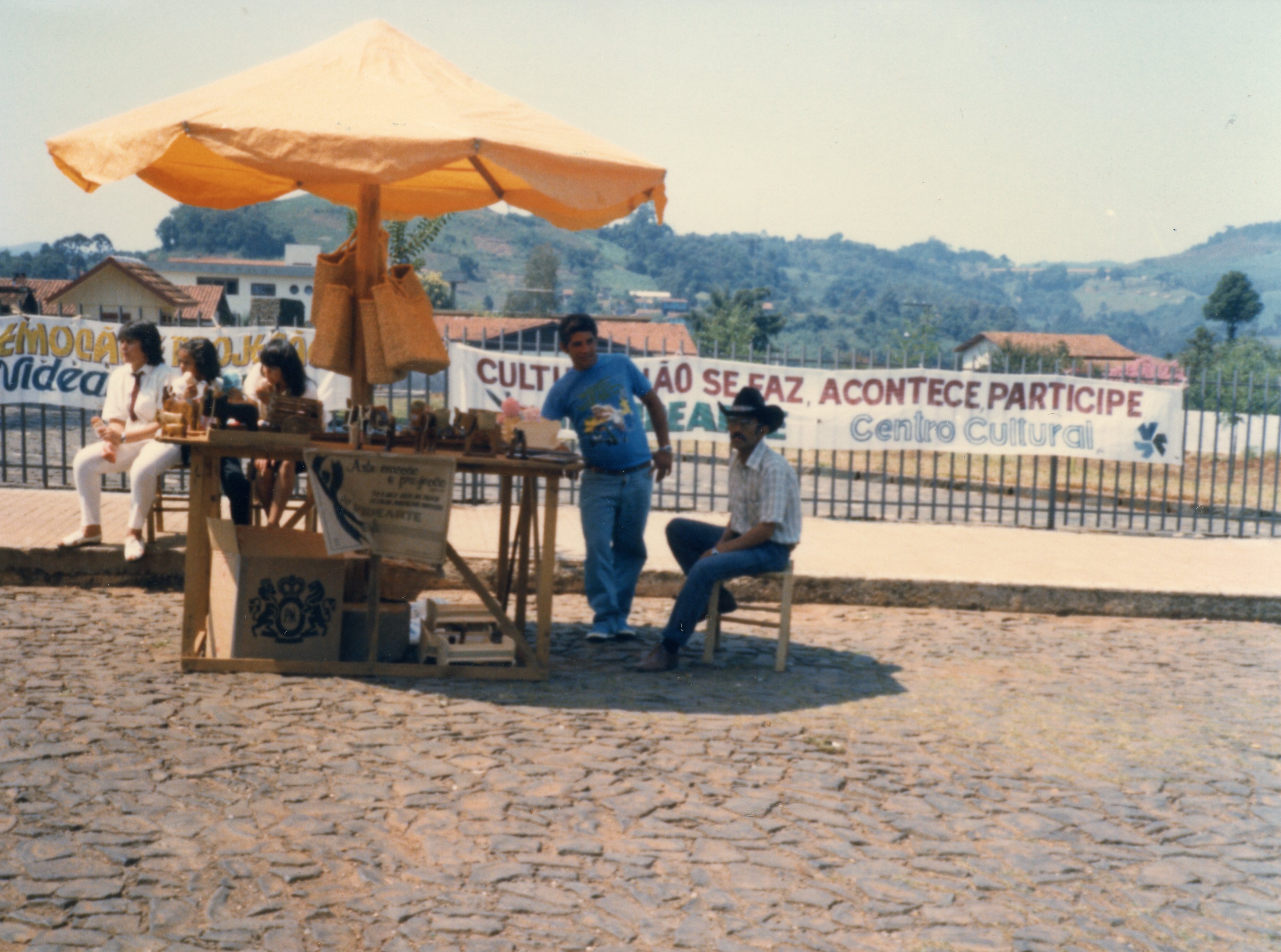 Uma cena ao ar livre em uma praça ou área pública, possivelmente em Videira, mostrando uma barraca de artesanato com guarda-sol amarelo. Pessoas estão sentadas e em pé ao redor da banca, com uma faixa ao fundo promovendo um evento do Centro Cultural, sugerindo uma feira de artesanato ou evento comunitário típico das décadas de 1980 ou 1990.