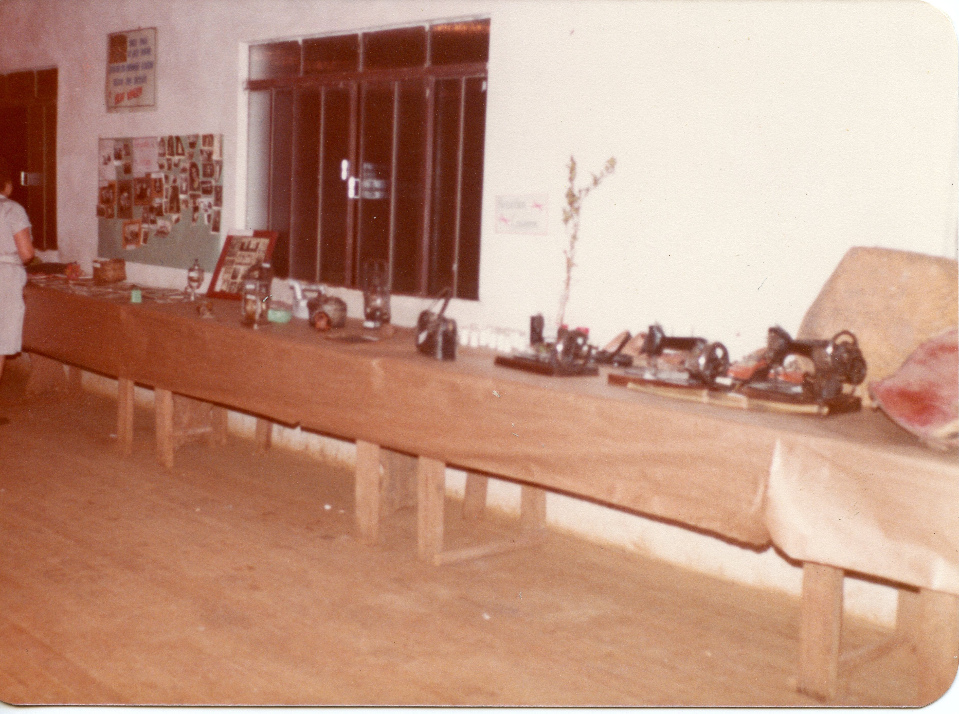 Uma fotografia interna que mostra uma exposição de objetos antigos, incluindo máquinas de costura manuais e itens domésticos dispostos sobre uma longa mesa de madeira. O ambiente parece ser uma sala comunitária ou escolar, com murais de fotos na parede, sugerindo uma exposição histórica ou cultural em uma pequena cidade brasileira.