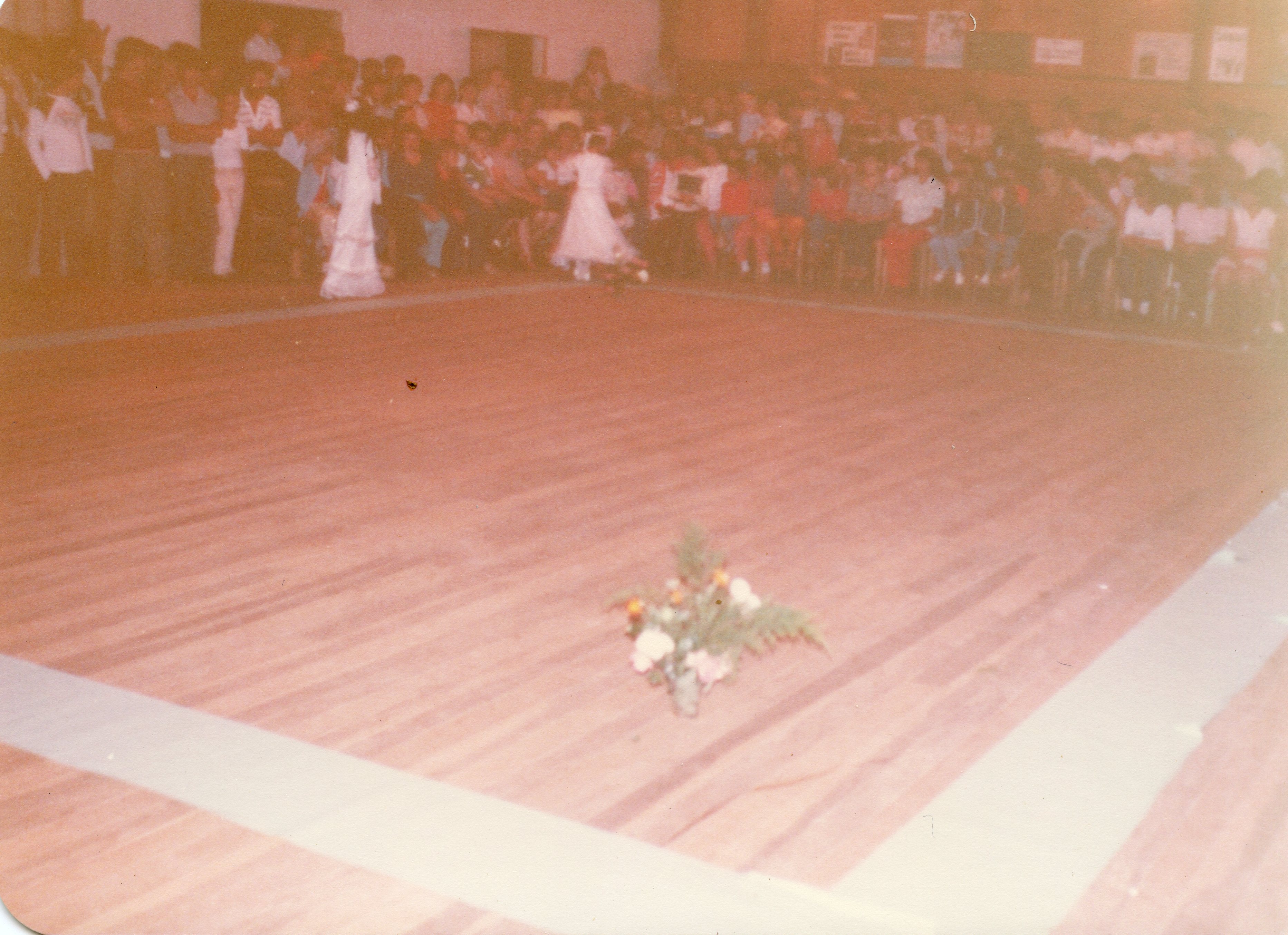 Uma fotografia de época, possivelmente das décadas de 1970 ou 1980, capturando um evento social ou escolar em um salão com piso de madeira. Observa-se uma grande plateia sentada ao fundo e uma criança vestida de branco no centro do salão, sugerindo uma apresentação ou cerimônia em uma comunidade brasileira.