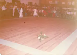 Uma fotografia de época, possivelmente das décadas de 1970 ou 1980, capturando um evento social ou escolar em um salão com piso de madeira. Observa-se uma grande plateia sentada ao fundo e uma criança vestida de branco no centro do salão, sugerindo uma apresentação ou cerimônia em uma comunidade brasileira.