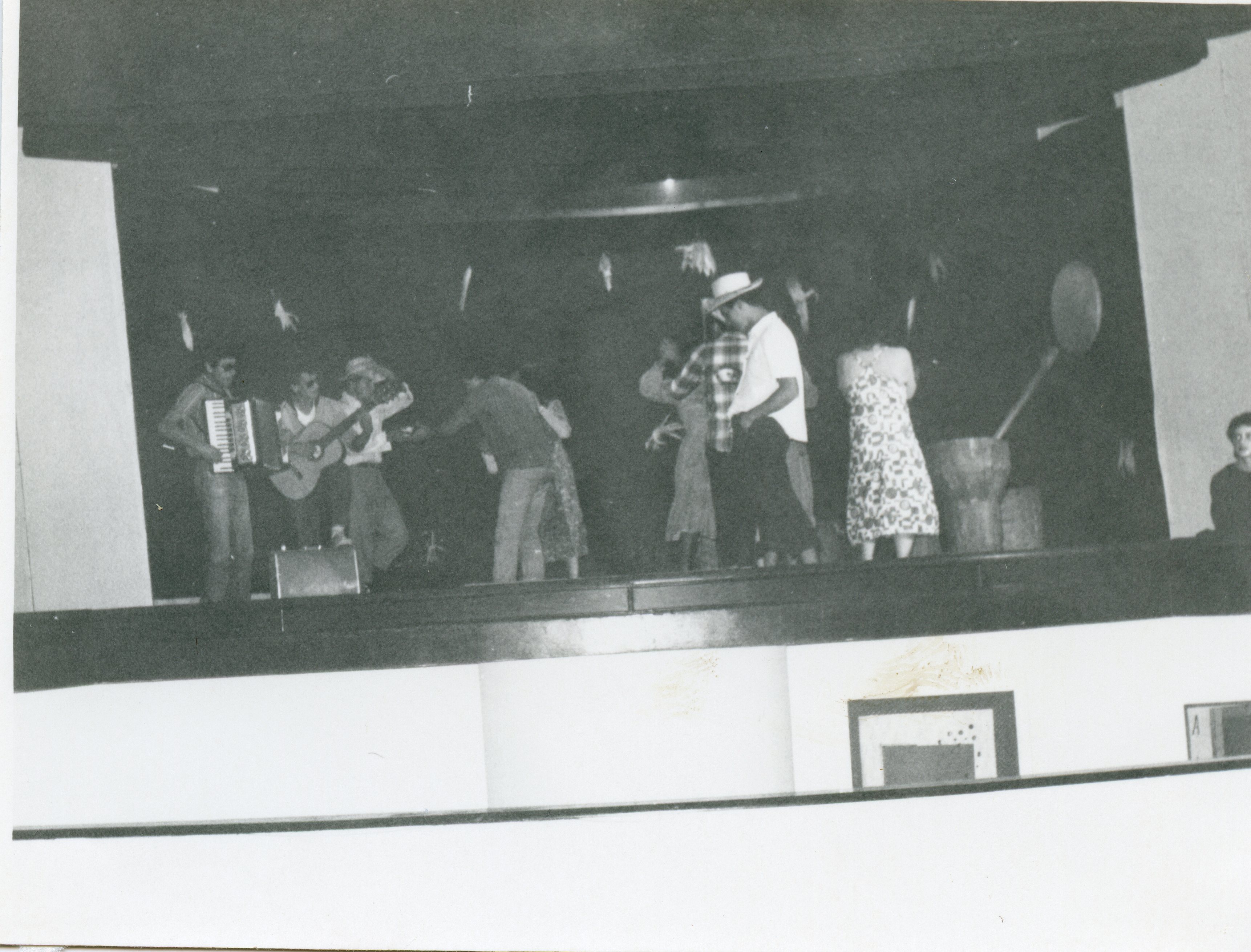 Uma fotografia em preto e branco que registra uma apresentação cultural ou festa típica em um palco, possivelmente uma festa junina devido aos trajes e elementos decorativos. O grupo inclui músicos com sanfona e violão, além de pessoas dançando, sugerindo um evento comunitário ou escolar de meados do século XX.