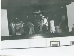 Uma fotografia em preto e branco que registra uma apresentação cultural ou festa típica em um palco, possivelmente uma festa junina devido aos trajes e elementos decorativos. O grupo inclui músicos com sanfona e violão, além de pessoas dançando, sugerindo um evento comunitário ou escolar de meados do século XX.