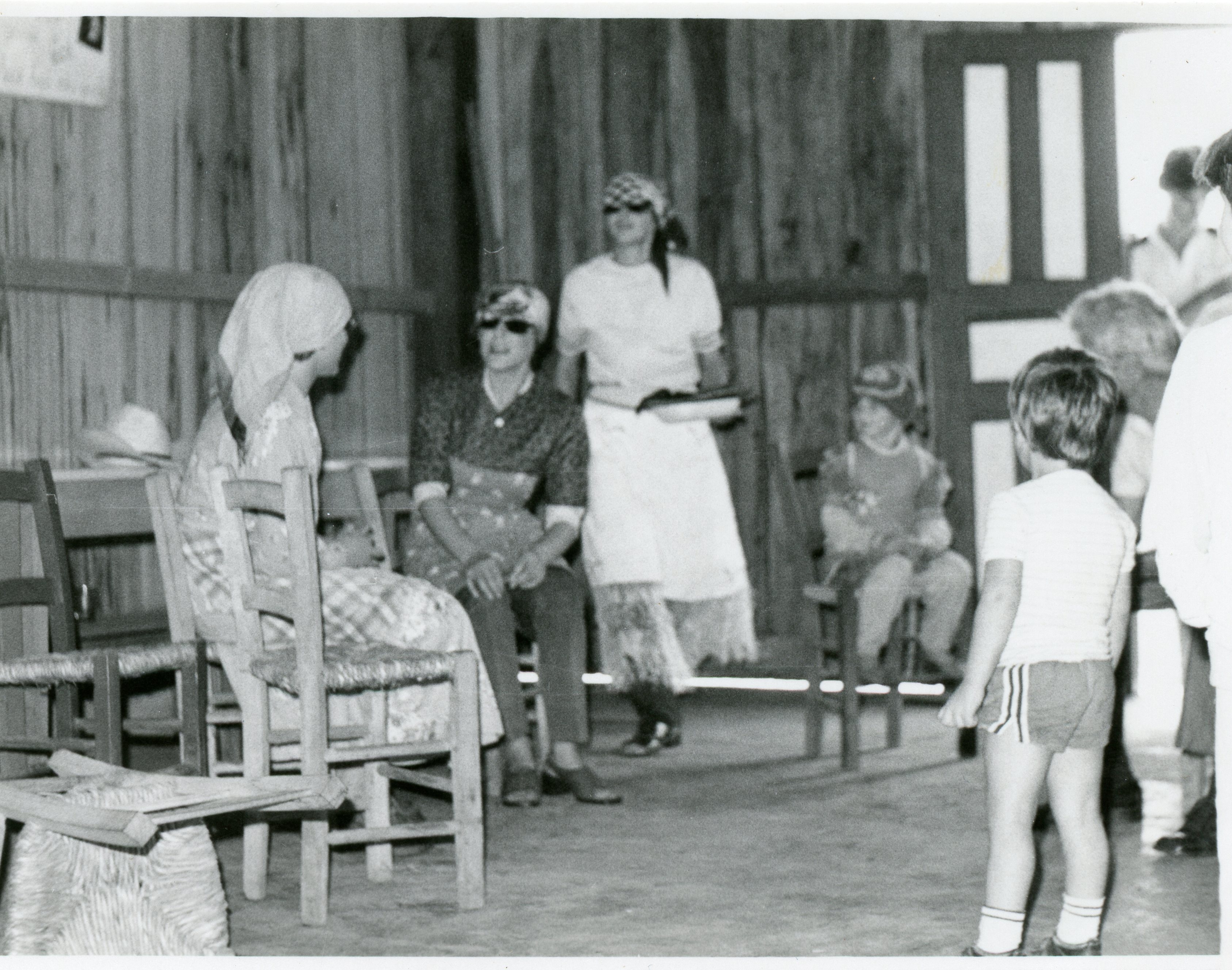 Fotografia em preto e branco registrando um encontro informal em um ambiente interno com paredes de madeira, possivelmente uma residência rural ou salão comunitário. Mulheres vestidas com aventais e lenços na cabeça conversam enquanto uma criança observa a cena, sugerindo um contexto de vida cotidiana ou evento comunitário de meados do século XX.
