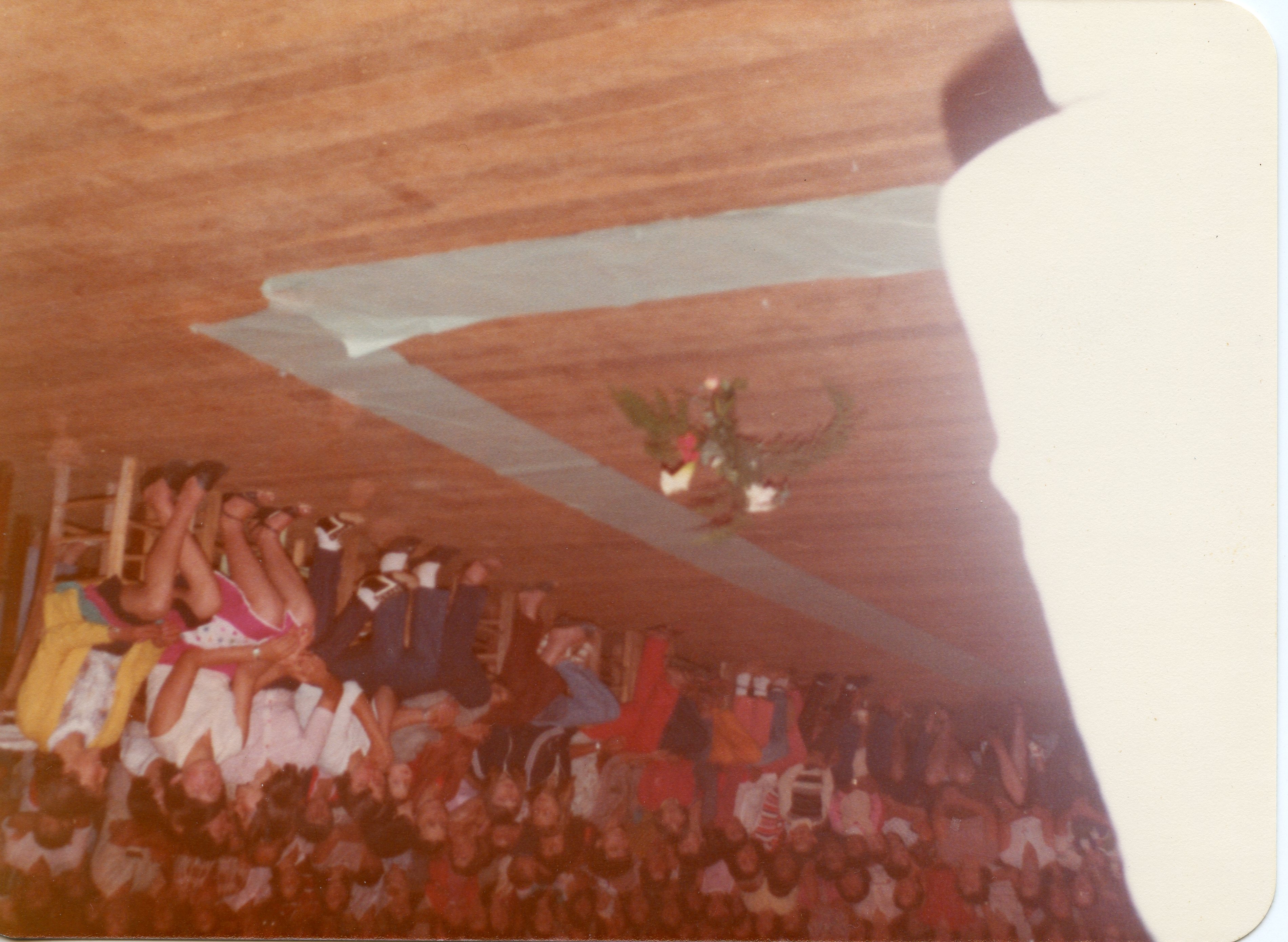 Uma fotografia colorida, orientada verticalmente, que captura um grupo de pessoas sentadas em cadeiras de madeira em um ambiente interno, possivelmente um salão comunitário ou igreja. A imagem apresenta um arranjo floral centralizado no chão e uma decoração simples, sugerindo um evento social ou religioso de meados da década de 1970 ou 1980.