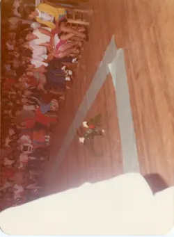 Uma fotografia colorida, orientada verticalmente, que captura um grupo de pessoas sentadas em cadeiras de madeira em um ambiente interno, possivelmente um salão comunitário ou igreja. A imagem apresenta um arranjo floral centralizado no chão e uma decoração simples, sugerindo um evento social ou religioso de meados da década de 1970 ou 1980.