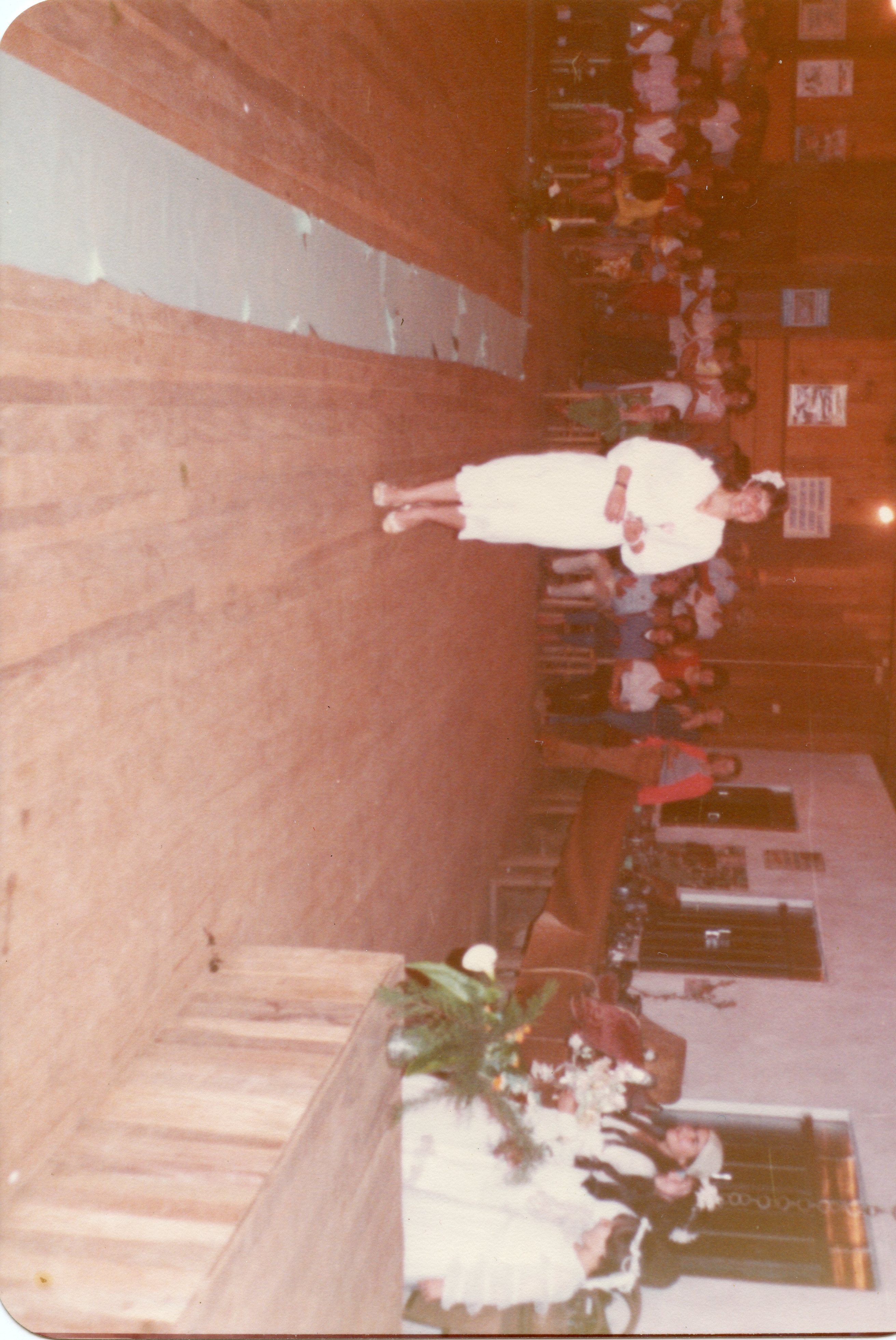 Uma fotografia de um evento social, possivelmente uma cerimônia religiosa ou escolar, realizada em um salão com piso de madeira. Vê-se uma pessoa vestida de branco caminhando pelo salão, enquanto outras pessoas observam sentadas ou em pé ao fundo, sugerindo uma celebração comunitária típica de meados da década de 1980.