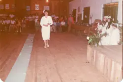 Uma fotografia de um evento social, possivelmente uma cerimônia religiosa ou escolar, realizada em um salão com piso de madeira. Vê-se uma pessoa vestida de branco caminhando pelo salão, enquanto outras pessoas observam sentadas ou em pé ao fundo, sugerindo uma celebração comunitária típica de meados da década de 1980.