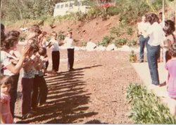 Fotografia colorida de época, possivelmente das décadas de 1970 ou 1980, retratando um grupo de pessoas em um ambiente externo, possivelmente em uma área rural ou de lazer. As vestimentas e a qualidade da imagem sugerem um registro casual ou familiar, com o grupo posicionado em uma encosta ou terreno inclinado.