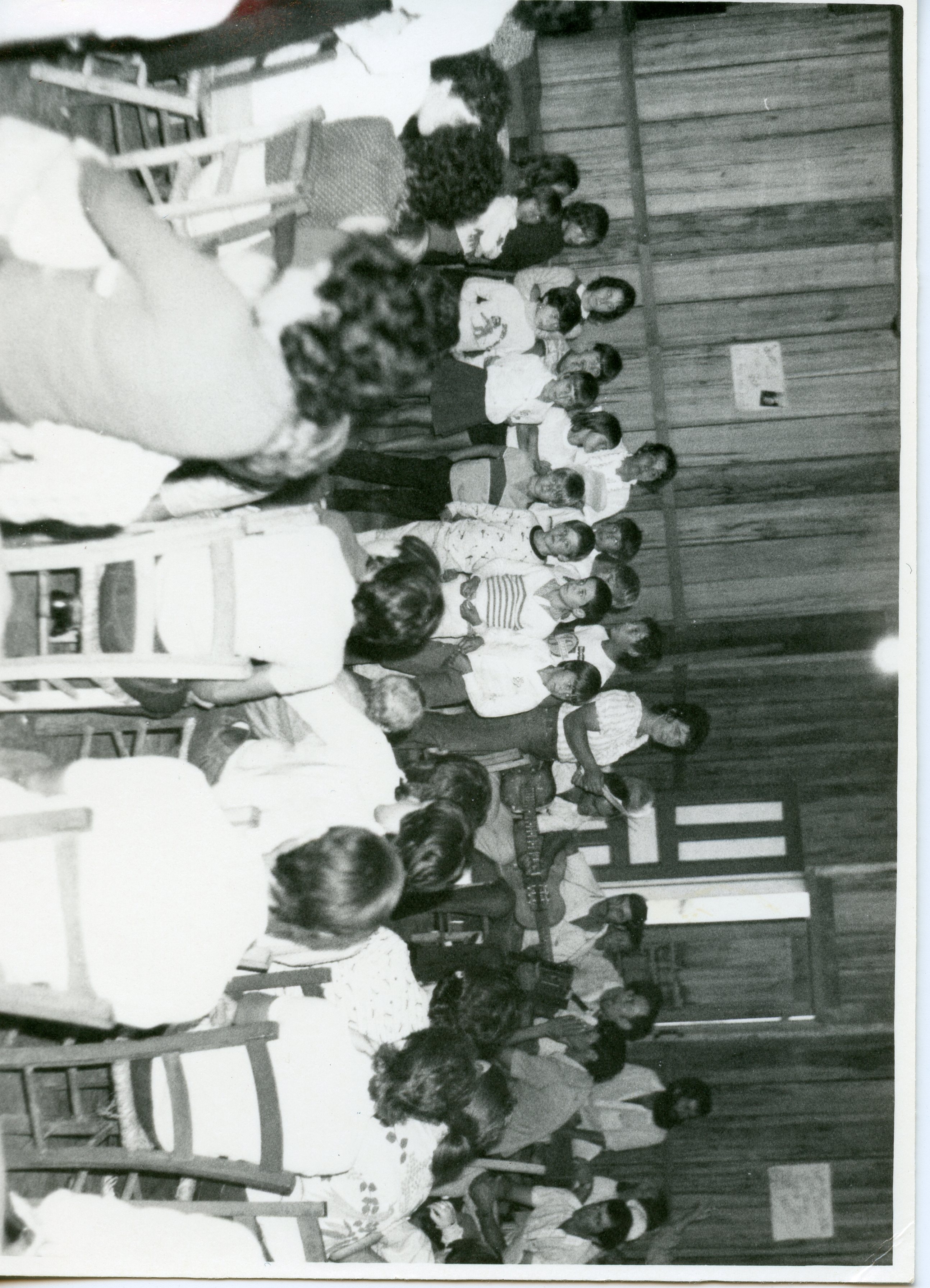 Fotografia em preto e branco que registra uma reunião comunitária ou escolar em um ambiente interno com paredes de madeira. O grupo, composto por diversas pessoas, parece estar participando de uma atividade social ou cultural, possivelmente em uma área rural ou pequena cidade brasileira, sugerindo um contexto de meados do século XX.
