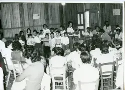 Fotografia em preto e branco que registra uma reunião comunitária ou escolar em um ambiente interno com paredes de madeira. O grupo, composto por diversas pessoas, parece estar participando de uma atividade social ou cultural, possivelmente em uma área rural ou pequena cidade brasileira, sugerindo um contexto de meados do século XX.