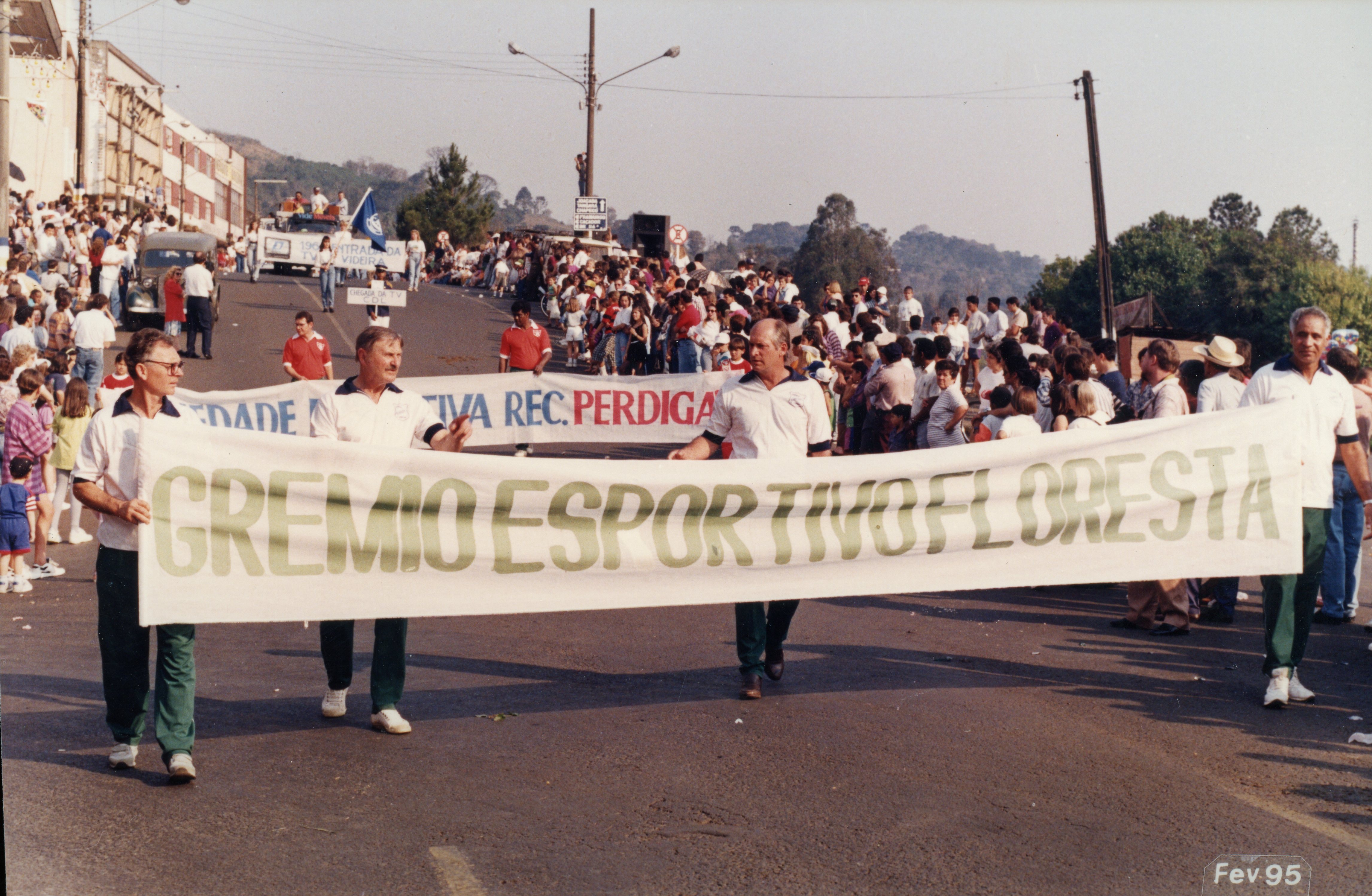 Uma fotografia de um desfile ou evento público em uma rua, onde homens carregam uma faixa do Grêmio Esportivo Floresta. O contexto sugere uma celebração comunitária ou esportiva em uma cidade brasileira, com a presença de público nas calçadas e veículos ao fundo, datada de fevereiro de 1995.