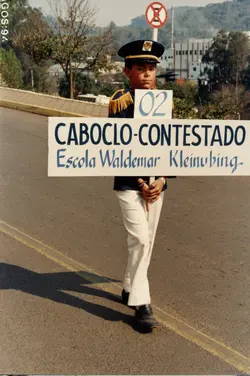 Uma criança vestida com uniforme de gala escolar segura uma placa durante um desfile cívico, possivelmente em Videira, Santa Catarina. A placa faz referência ao tema 'Caboclo-Contestado' e à Escola Waldemar Kleinubing, sugerindo um evento comemorativo ou histórico da década de 1990.
