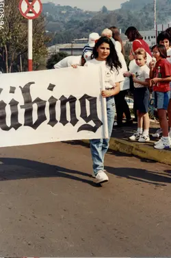 Uma fotografia colorida que registra um grupo de pessoas, possivelmente em um desfile ou evento comunitário, carregando uma faixa com a inscrição 'Dübing'. A cena apresenta vestimentas típicas da década de 1990, como calças jeans de cintura alta e camisetas casuais, em um ambiente urbano ao ar livre.