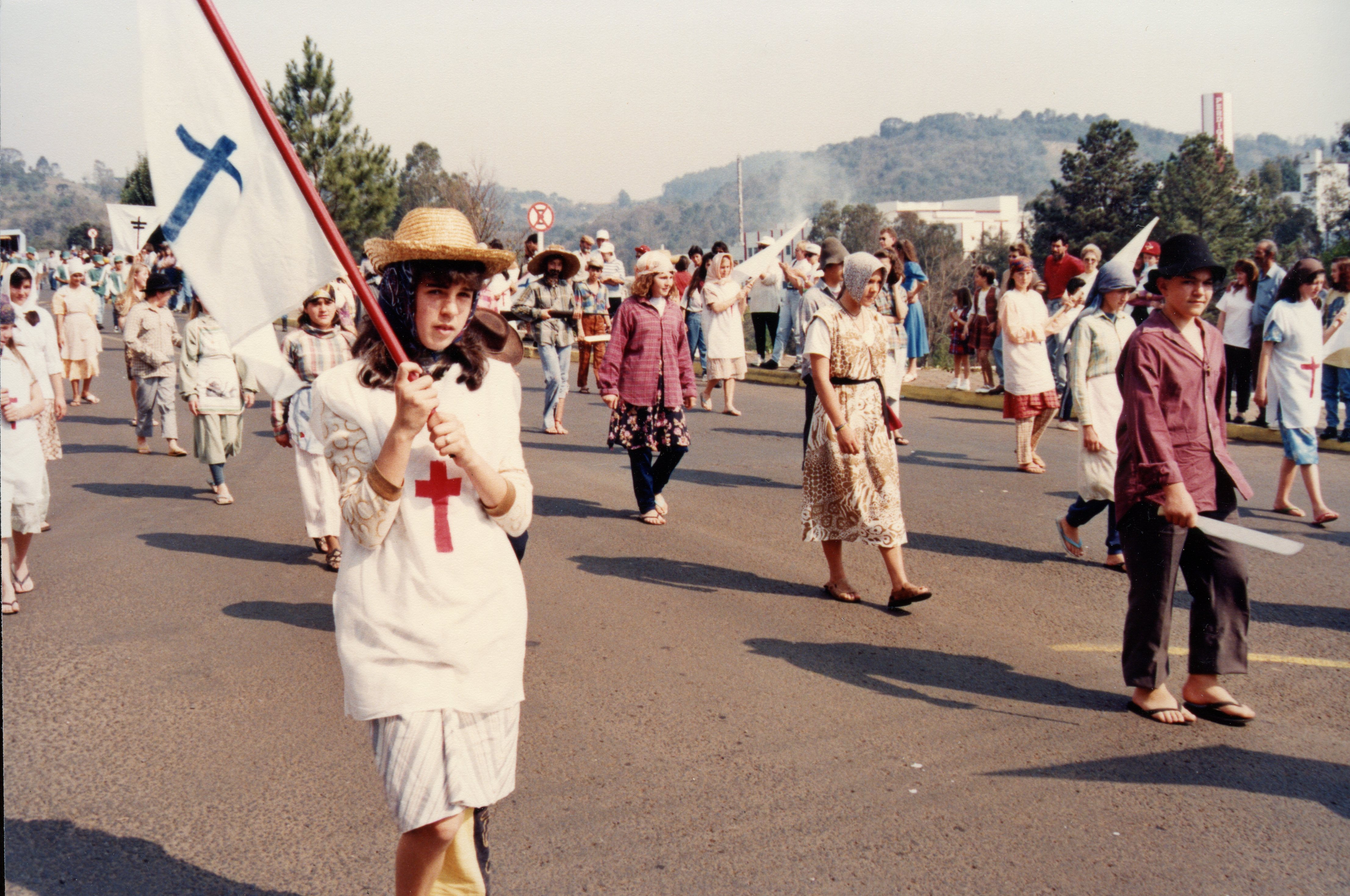 Uma procissão ou desfile cívico-religioso ao ar livre, onde jovens carregam bandeiras com símbolos cristãos, como cruzes. A cena sugere uma celebração comunitária em uma cidade brasileira, possivelmente nas décadas de 1980 ou 1990, com participantes vestindo roupas casuais da época.