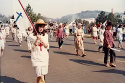 Uma procissão ou desfile cívico-religioso ao ar livre, onde jovens carregam bandeiras com símbolos cristãos, como cruzes. A cena sugere uma celebração comunitária em uma cidade brasileira, possivelmente nas décadas de 1980 ou 1990, com participantes vestindo roupas casuais da época.