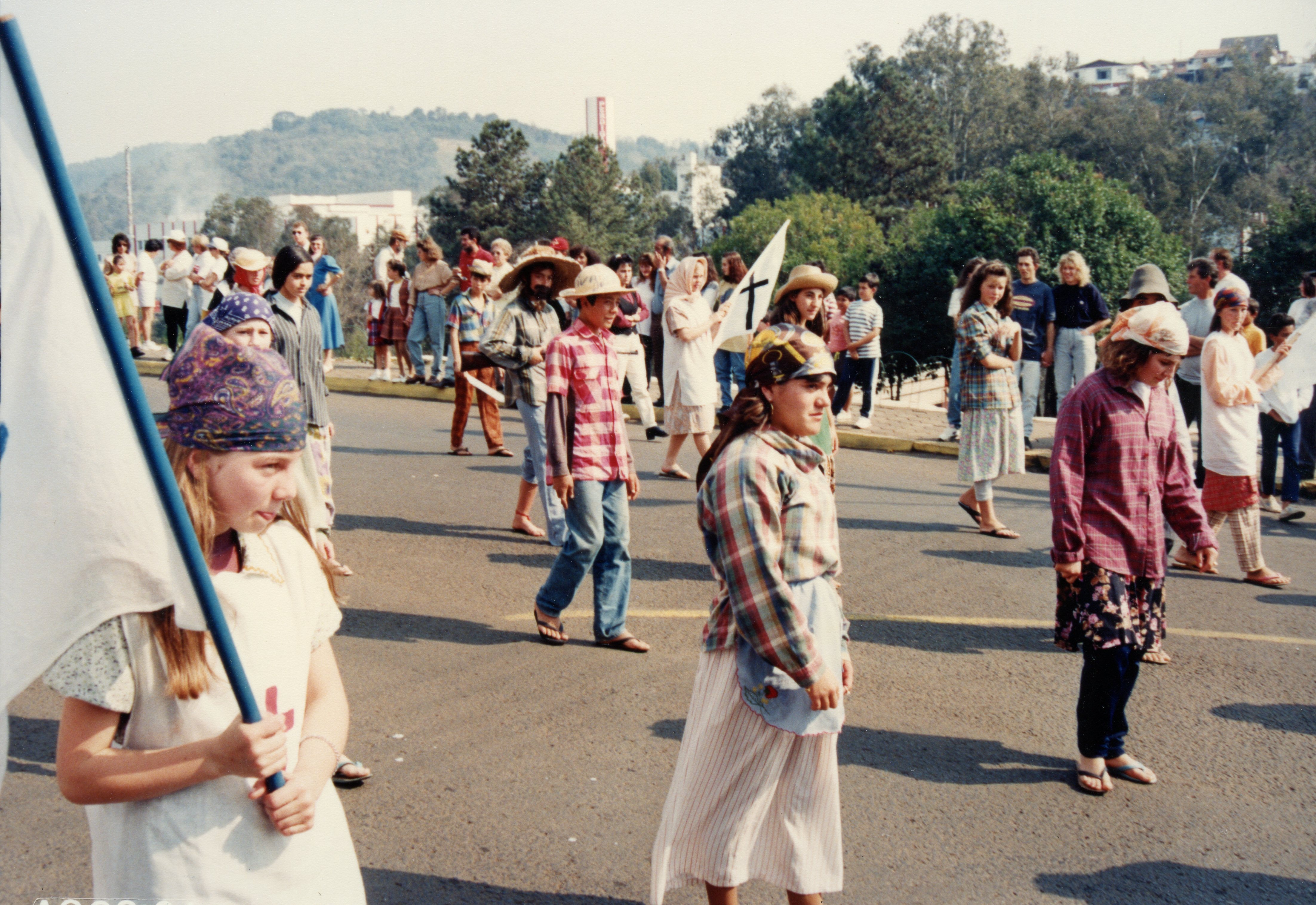 Uma cena de desfile ou procissão ao ar livre, possivelmente um evento escolar ou comunitário, com jovens vestindo trajes rústicos e chapéus. O contexto sugere uma celebração cultural ou religiosa em uma cidade brasileira, com participantes carregando bandeiras e caminhando por uma via pavimentada.