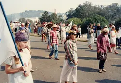 Uma cena de desfile ou procissão ao ar livre, possivelmente um evento escolar ou comunitário, com jovens vestindo trajes rústicos e chapéus. O contexto sugere uma celebração cultural ou religiosa em uma cidade brasileira, com participantes carregando bandeiras e caminhando por uma via pavimentada.