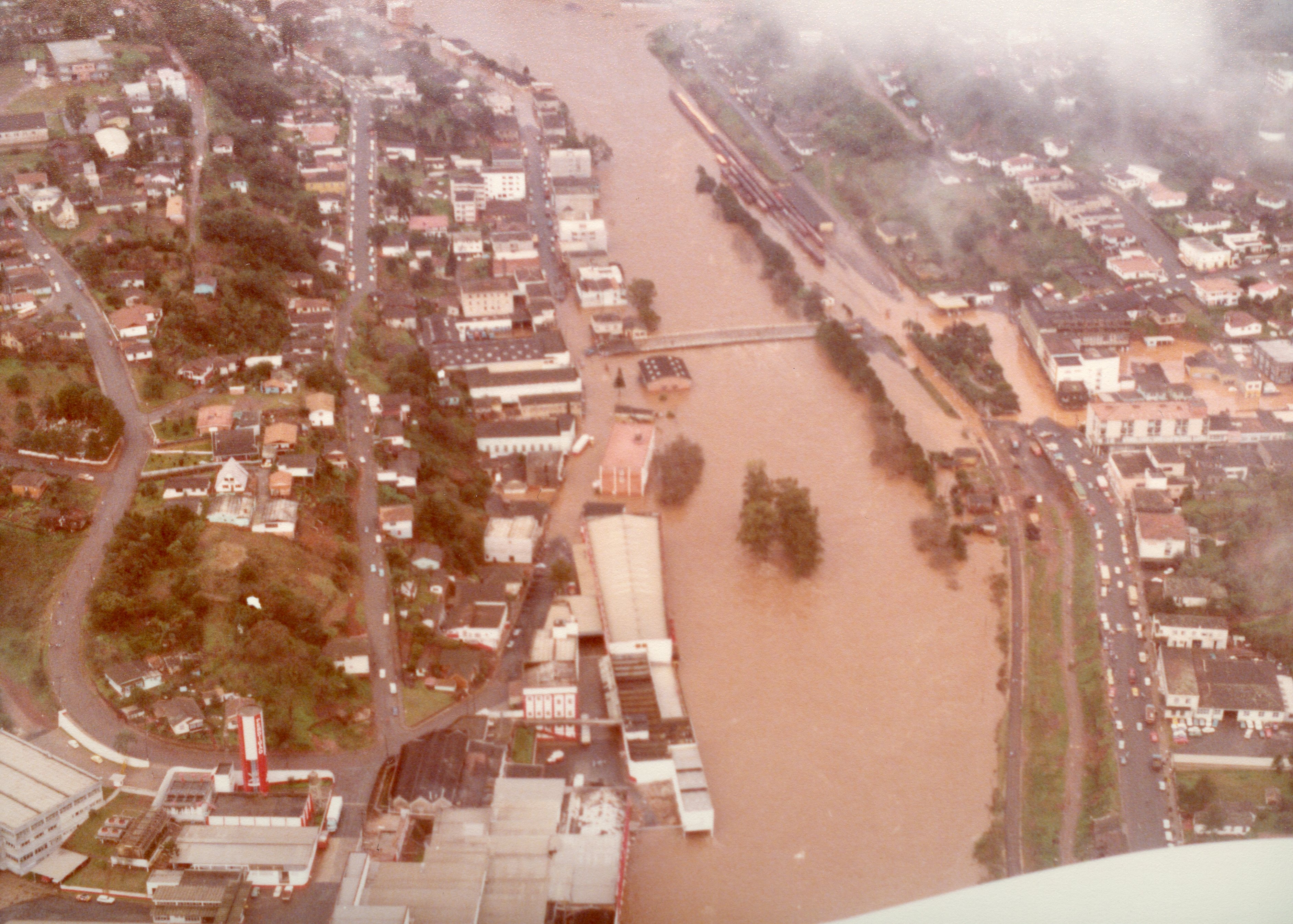 Uma vista aérea de uma enchente severa em uma cidade brasileira, possivelmente Videira, mostrando o rio transbordado cobrindo áreas urbanas e industriais. A imagem apresenta características de fotografia colorida de meados da década de 1980, com tons desbotados e uma perspectiva panorâmica que destaca o impacto do desastre natural na infraestrutura local.
