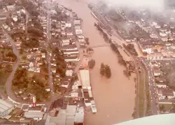 Uma vista aérea de uma enchente severa em uma cidade brasileira, possivelmente Videira, mostrando o rio transbordado cobrindo áreas urbanas e industriais. A imagem apresenta características de fotografia colorida de meados da década de 1980, com tons desbotados e uma perspectiva panorâmica que destaca o impacto do desastre natural na infraestrutura local.