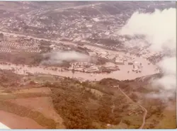 Uma fotografia aérea de uma enchente em uma área urbana, possivelmente em uma cidade do interior brasileiro. A imagem mostra um rio transbordado ocupando grande parte do vale, com construções próximas à margem submersas ou isoladas, sugerindo um evento climático severo de meados do século XX.