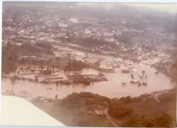 Fotografia aérea de uma enchente em uma área urbana, possivelmente em uma cidade do interior catarinense como Videira. A imagem mostra o rio transbordado cobrindo partes da infraestrutura urbana e industrial, com coloração barrenta característica de cheias.
