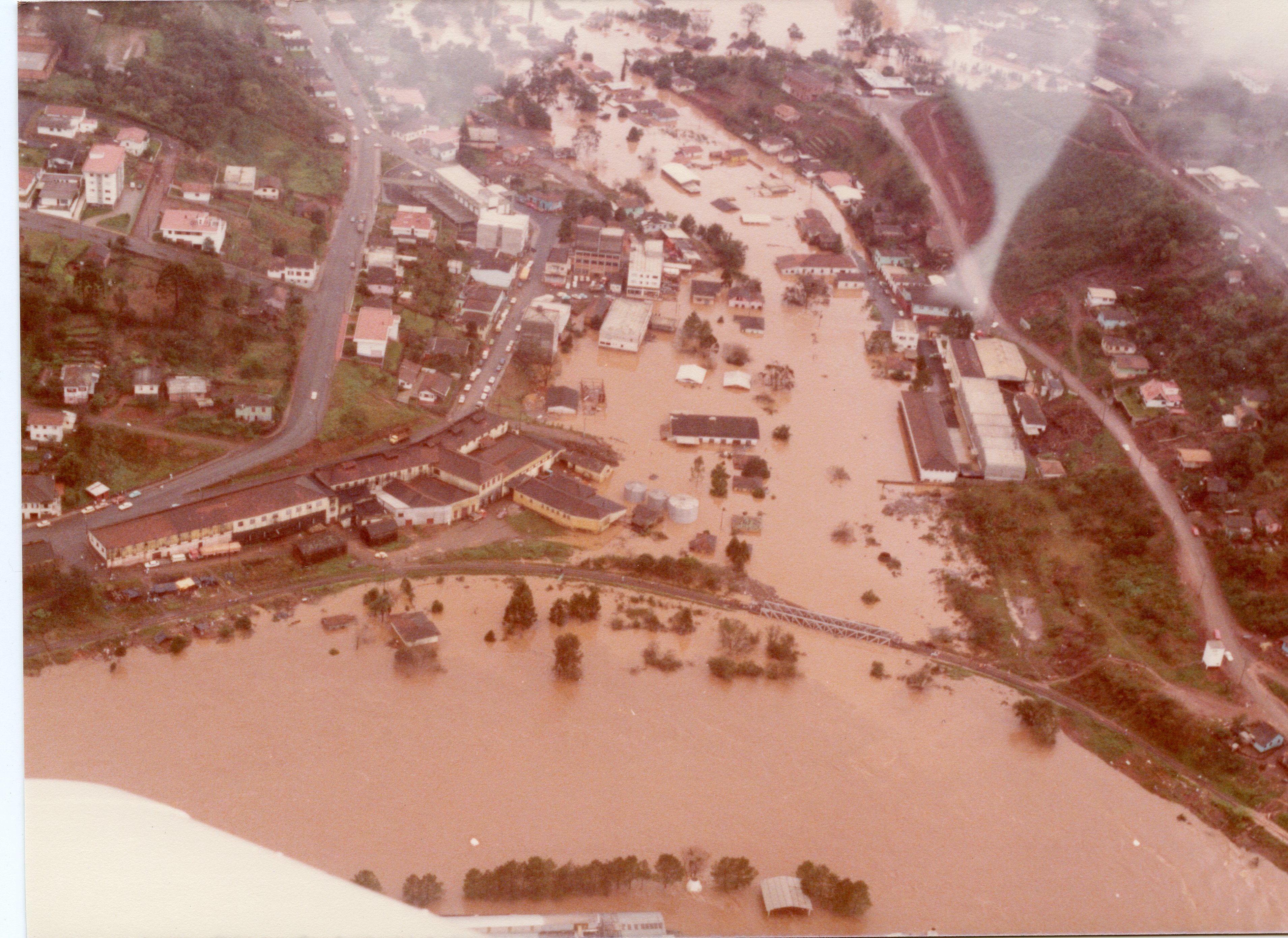 Fotografia aérea de uma enchente severa em uma área urbana, possivelmente em uma cidade do interior do sul do Brasil. A imagem mostra ruas e construções submersas por águas barrentas, com destaque para a infraestrutura urbana e uma ponte ferroviária parcialmente coberta, sugerindo um evento climático de grande impacto ocorrido provavelmente entre as décadas de 1970 e 1980.