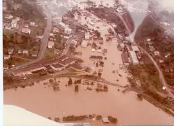 Fotografia aérea de uma enchente severa em uma área urbana, possivelmente em uma cidade do interior do sul do Brasil. A imagem mostra ruas e construções submersas por águas barrentas, com destaque para a infraestrutura urbana e uma ponte ferroviária parcialmente coberta, sugerindo um evento climático de grande impacto ocorrido provavelmente entre as décadas de 1970 e 1980.